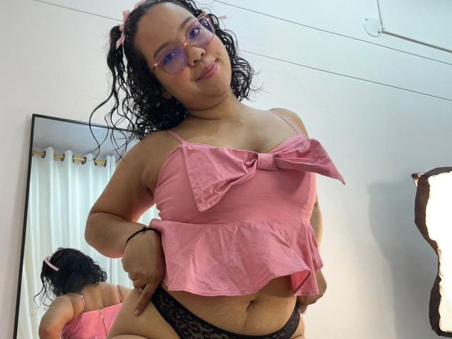 NicolleAry with curly black hair, pink bow, glasses, pink top, bow, showing her tits, black lace panties, hands on hips, mirror reflection, white curtain, soft smile.
