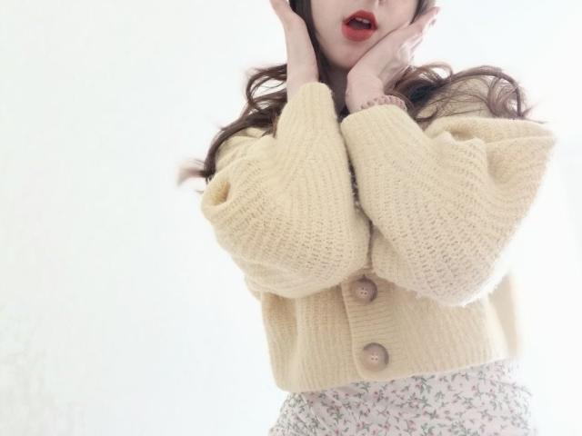 WollyMolly wearing a yellow knit sweater with buttons, holding her ears, red lips, curly brown hair, and floral skirt, hands up, face partially hidden.