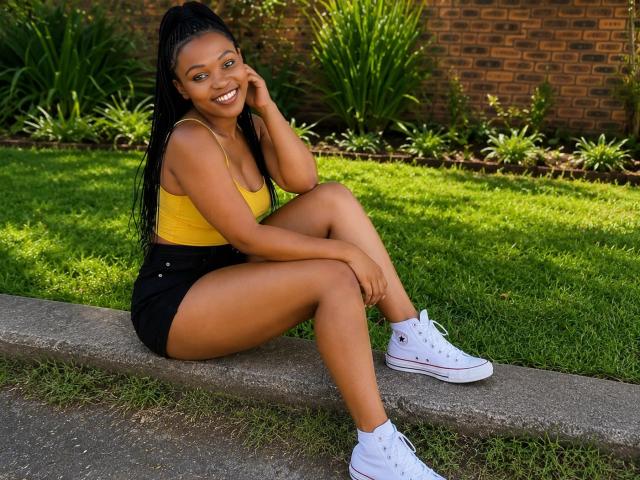 SpicyStorm with dark brown skin, long black hair in a ponytail, yellow tank top, black shorts, white sneakers, smiling, sitting on a concrete edge, green grass in background.
