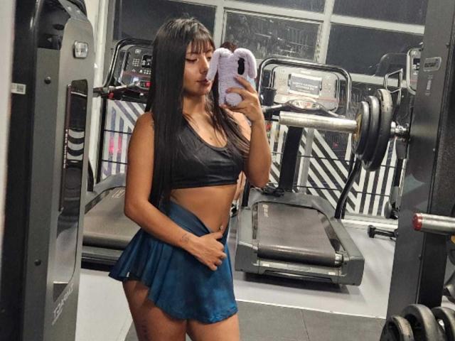 LeiaLaurent in a gym, wearing a black sports bra and blue skirt, holding a phone with a purple case, showing her taut abs and curvy ass.
