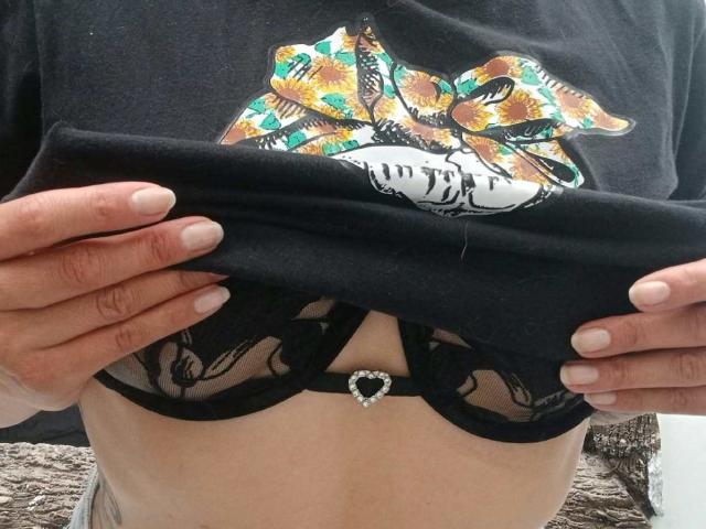 ZarayFeet solleva una maglietta nera, mostrando un reggiseno nero in pizzo con un centro a forma di cuore, rivelando i suoi seni. Mani con unghie bianche tengono la maglietta, pelle leggermente abbronzata..