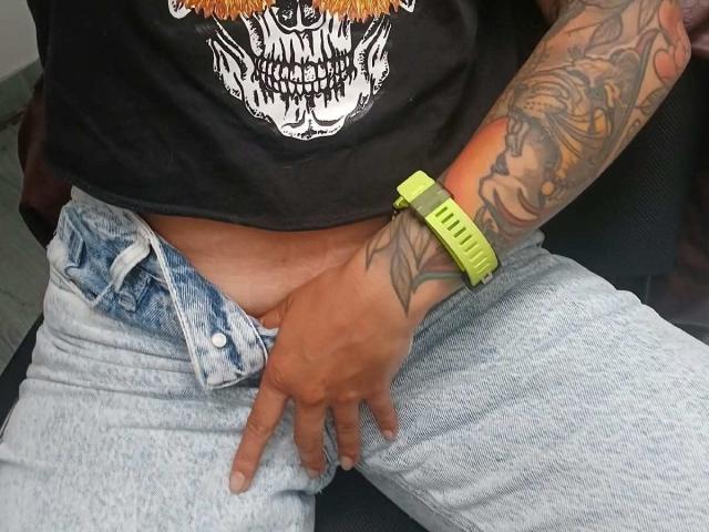 La mano di ZarayFeet, con il braccio tatuato, che indossa una maglietta nera con un teschio e un orologio verde neon, sta toccando i suoi jeans di denim chiaro, sbottonati, che scoprono la sua vulva, dalla pelle marrone e ricoperta di peli..