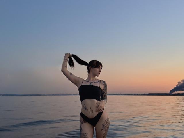 MissTattoo com biquíni preto, segurando o rabo de cavalo, tatuagens nos braços e pernas, em pé na água ao pôr do sol, céu azul a laranja, reflexos na água, mão direita no quadril..