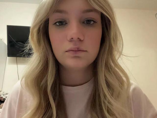 TrishGenna tiene el pelo rubio largo, piel clara y lleva una camisa rosa. Mira directamente a la cámara con una expresión neutra. Tiene una nariz pequeña, poco maquillaje y ojos azules..