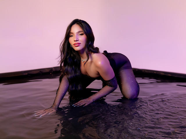 KatheVera with long black hair, wearing a black mesh bodysuit and gloves, on all fours in a black water-filled room, showing her tits and ass, with a pinkish light.