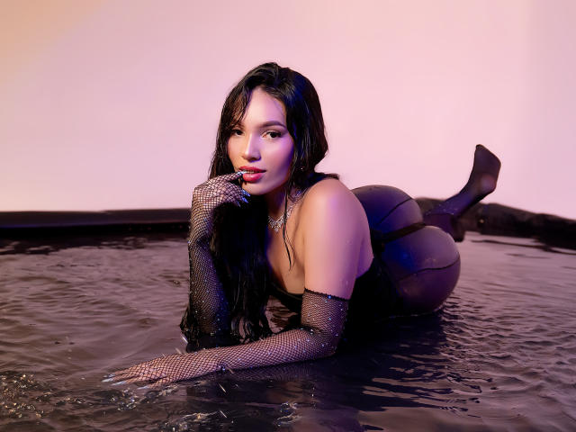 KatheVera with long black hair, wearing fishnet gloves and black lingerie, lying on her stomach in water, showing her ass and red lips, with a seductive expression.