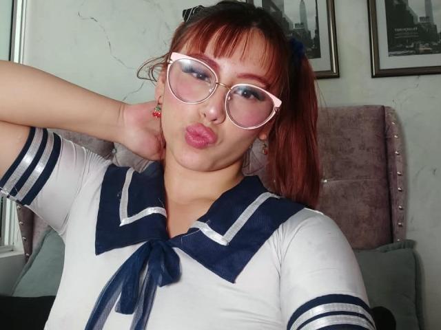 MaddyMills mit dicken, rosa Brillen, roten Zöpfen und einem japanischen Schulmädchen-Outfit, die ihre Lippen zusammenpresst. Sie zeigt ihre Brüste, wobei ein Arm hinter ihrem Kopf ist..