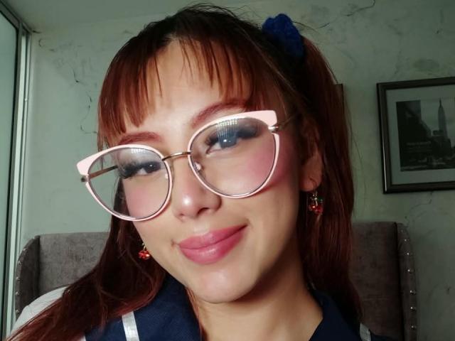 MaddyMills mit randlosen rosa Brillen, rotem Lippenstift und braunen Zöpfen, die einen blauen Haargummi trägt, lächelnd, mit roten Ohrringen, und sitzend auf einem Stuhl..