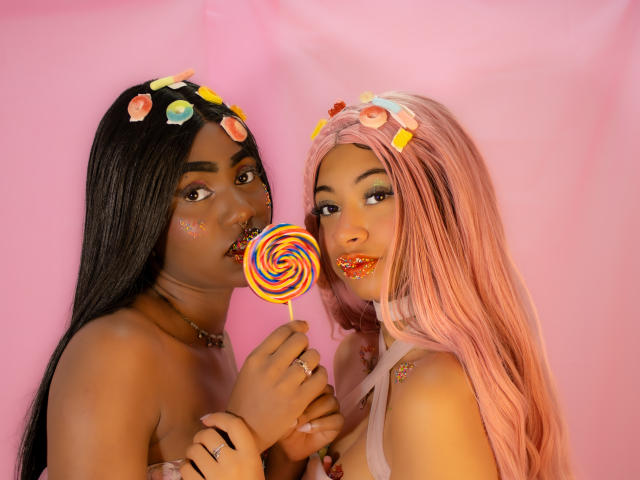 HaryAndKimmy with long black hair and pink hair holding a rainbow swirl candy, pink lipstick, flower headbands, and looking at camera, pink background, cute, sexy, colorful.