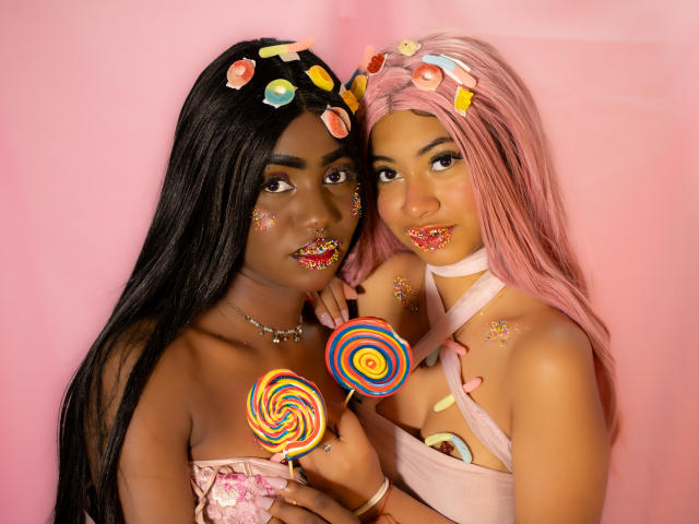 HaryAndKimmy, two dark-skinned women with long black and pink hair, holding colorful swirl lollipops, face smeared with candy, wearing strapless dresses, pink background.