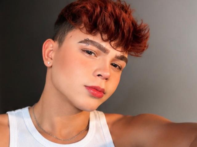 O PrinceSteve tem cabelo curto, ruivo e encaracolado, olhos castanhos, um piercing de prata no nariz e uma camiseta branca. Seus lábios são vermelhos e ele está usando um colar de prata fino..