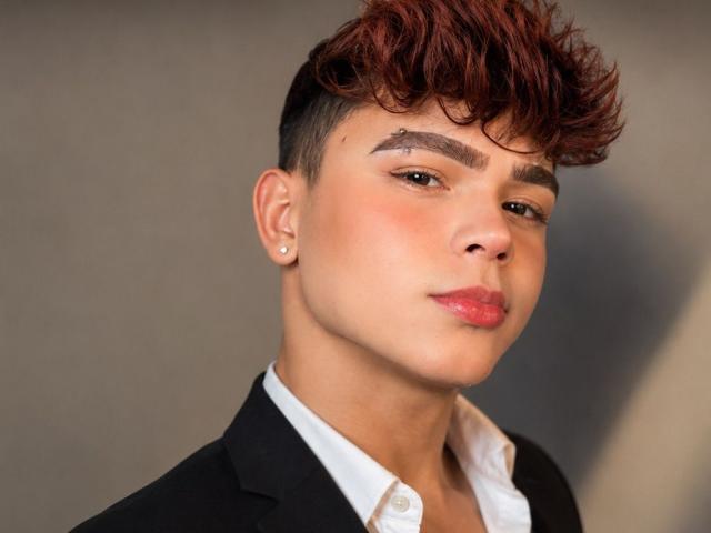 PrinceSteve con capelli castano corti, riflessi rossi, sopracciglia scure, orecchio con piercing, camicia bianca, giacca nera, labbra rosa, leggero sorriso, che guarda la fotocamera, illuminazione soffusa, pelle liscia..