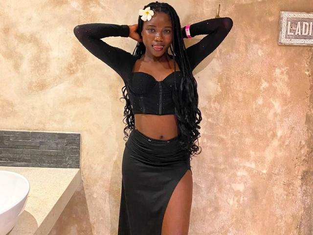 EbonySweet-hot in a black sparkly crop top and high-slit black skirt, hands behind head, long braids, white flower in hair, smiling, standing near a bathroom sink.