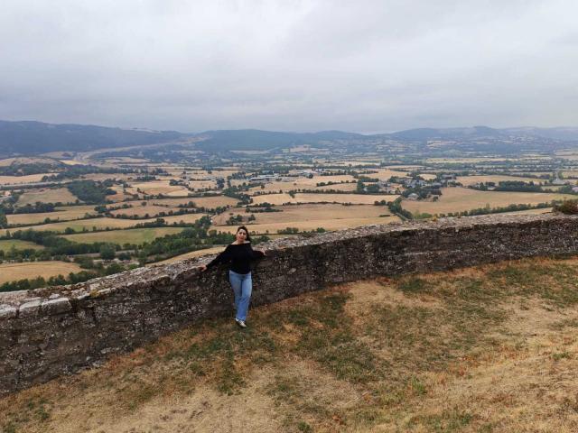 EtoileLatin leans against a stone wall, wearing a black top and blue jeans, with her back to the camera, on a grassy, slightly muddy hill.