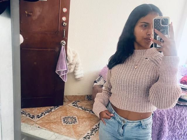 MyaaTyler with long black hair, wearing a pink knit sweater and blue jeans, holding a phone in a mirror selfie, showing her small tits and slim waist.