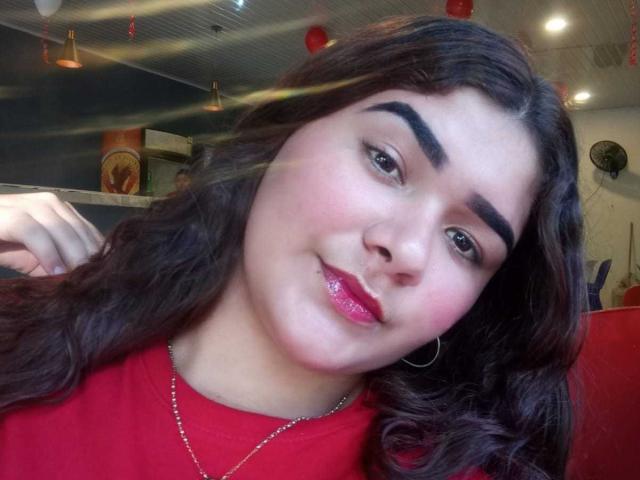 Zoe Estrada tem cabelos castanhos cacheados, sobrancelhas grossas, lábios rosados e brilhantes, e uma camisa vermelha, usando brincos de argola e um colar de prata, com um sorriso discreto e um rosto adorável..