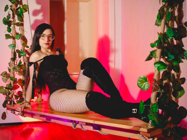 JadeHarpper with long dark hair, glasses, black off-shoulder top, fishnet stockings, and black thigh-high boots, sitting on a wooden swing with green leaf vines. Red light.
