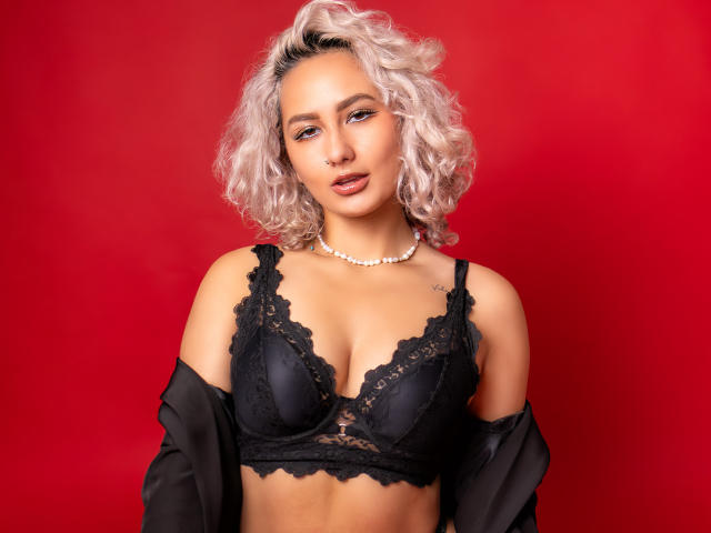 ElenaSaenz with curly pink hair, wearing black lace bra, pearl necklace, and black sheer robe, showing her tits against a red background. She has a tattoo on her right shoulder.