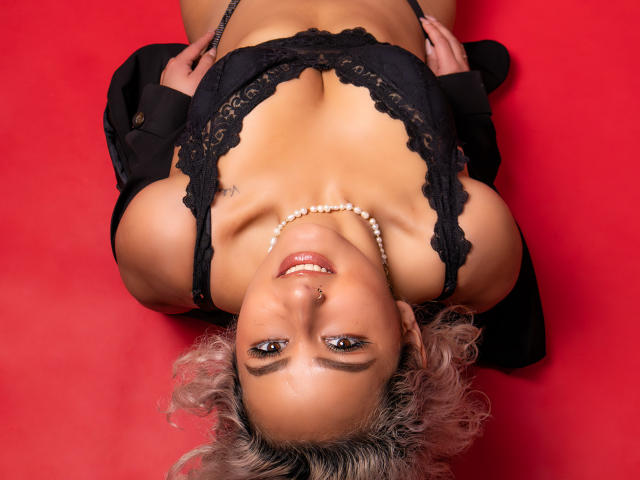 ElenaSaenz, lying on red, wearing black lace top, pearl necklace, black jacket, smiling, nose ring, curly blonde hair, hands on chest, medium tits, visible cleavage, attractive.