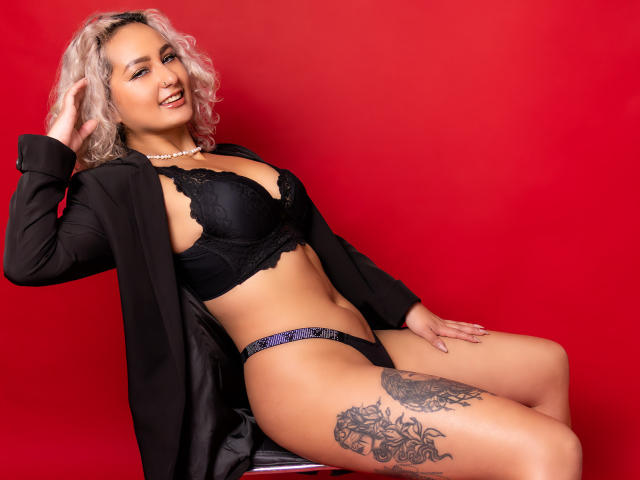 ElenaSaenz with blonde curly hair, black lace bra, black panties, black blazer, and pearl necklace, smiling, tattoo on right thigh, sitting on chair against red background.