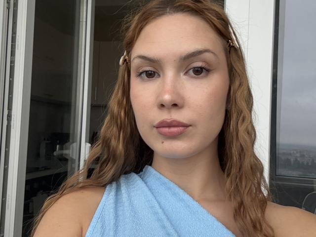 RebbecaNeelon has wavy brown hair, brown eyes, and a small mole on her chin. She's wearing a blue towel over one shoulder, with hair clips on both sides.