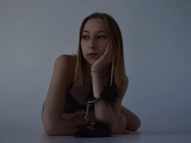 KleoYork with blonde hair, black bra, and black fluffy wrist cuffs, lying down, hand on cheek, looking to the side, in dim light, chains on wrist cuff.
