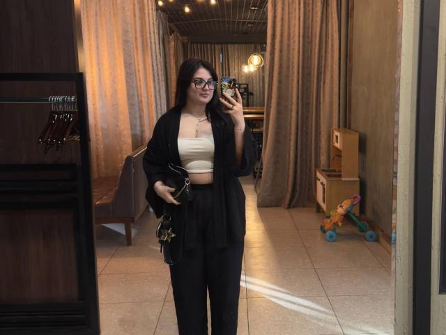 LisaMacdougall with glasses, black robe, white crop top, black pants, holding phone, standing in room with mirror, brown curtains, and wooden shelves, showing her ass.