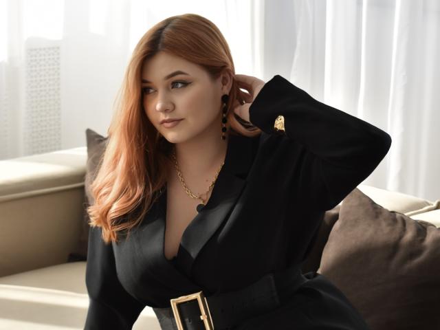 AmySweete with orange hair, black dress, gold chain, and earrings, hand in hair, sitting on beige couch, looking slightly to the side, soft light, subtle smile.