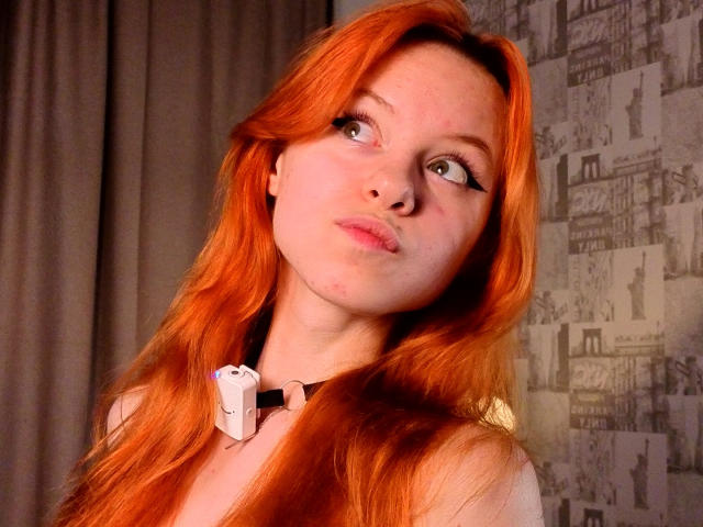 JoiMazey has long, orange-red hair, pale skin, and green eyes, wearing a white headset with a black strap, looking up with a pouty, slightly freckled face.