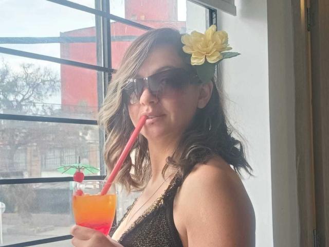 ArianaBlonde with wavy brown hair, wearing sunglasses and a yellow flower in her hair, holding a colorful drink with a pink straw, showing her tits in a brown top.