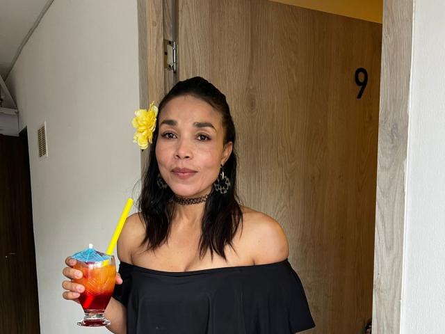 MiaLucchi with medium tits, wearing a black off-shoulder top, choker, and yellow flower in hair, holding a colorful drink with a yellow straw, smiling.