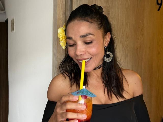 MiaLucchi with dark hair in a ponytail, yellow flower, black off-shoulder top, chomping on a straw from a fruity drink with a paper umbrella, smiling.