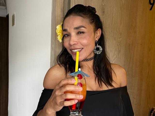 MiaLucchi with dark hair, yellow flower, black choker, and off-shoulder black top, sipping a drink with a yellow straw, red cherry, and blue umbrella, smiling.