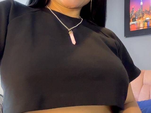 CharlotteCrofort's black crop top, showing her big tits, with a pink crystal necklace and black hair. Her face is partially visible.