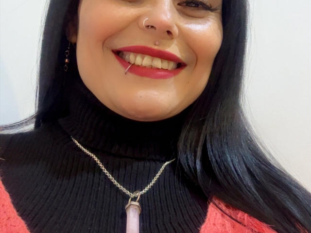 CharlotteCrofort has long black hair, red lipstick, a nose ring, and a toothpick in her mouth. She's wearing a red and black turtleneck with a pink necklace.
