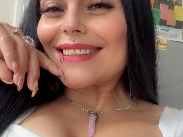CharlotteCrofort with black hair, pink lipstick, nose ring, and purple pendant, smiling with teeth showing, hand with long nails resting on cheek, white top, pink nails.