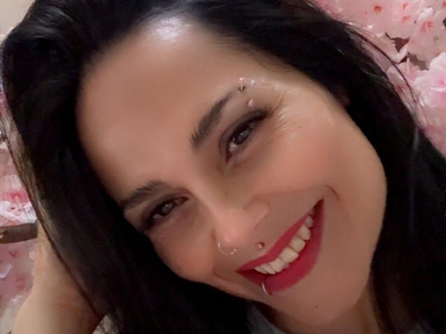 CharlotteCrofort has black hair, a nose ring, and a lip piercing, smiling with red lipstick, and has a small gold bar above her upper lip.