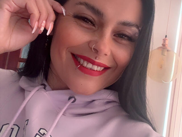 CharlotteCrofort with red lipstick, nose ring, and lip piercing, smiling at the camera, wearing a light purple hoodie, long black hair, and white nails.