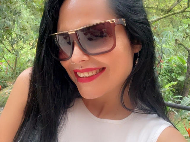 CharlotteCrofort with black hair, red lipstick, nose ring, white tank top, and large brown sunglasses, smiling outdoors. Her tits are not visible. She's standing in a green, leafy area.