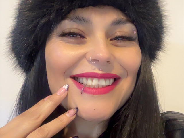 CharlotteCrofort with black fur hat, nose ring, and pink lipstick, smiling with one eye closed, finger touching her cheek, white nail polish with pink design, nosebleed on her lip.