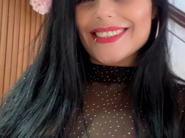 CharlotteCrofort with black hair, red lipstick, and a nose ring, smiling with a tooth partially visible, wearing a sheer black polka dot top, wooden background.