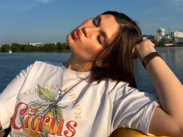 LakeishaCresencio with eyes closed, head tilted, brown hair, white t-shirt, "Californian" graphic, necklace, wristwatch, sitting by water, sunlight on face, hand in hair.