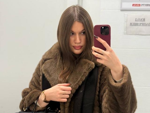 LakeishaCresencio with long brown hair, wearing a brown fur coat, taking a selfie with a pink phone, holding a black bag, wearing a gold watch, and ring.