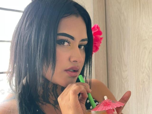 LairaHearvy with black hair, green eyeliner, and pink flower, applying yellow eyeshadow with green eyeliner brush, holding pink umbrella in her right hand, wood background.
