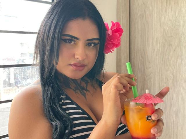LairaHearvy with dark hair, pink flower, and black-striped top, holding a colorful drink with a green straw and pink umbrella, showing her tits and tattooed neck.