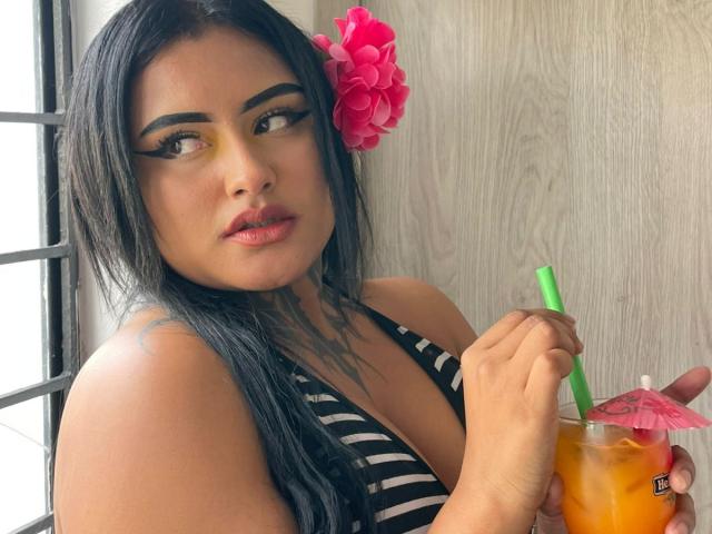 LairaHearvy with dark hair, pink flower in hair, bold eyeliner, and striped top, holding a drink with a pink umbrella and green straw, looking to the side.
