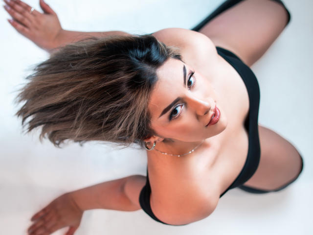 GabrielaMonique, with brown, messy hair, looking up, wearing black lingerie, has visible tits, a cute nose, and is on her knees, with legs spread, on a white floor.