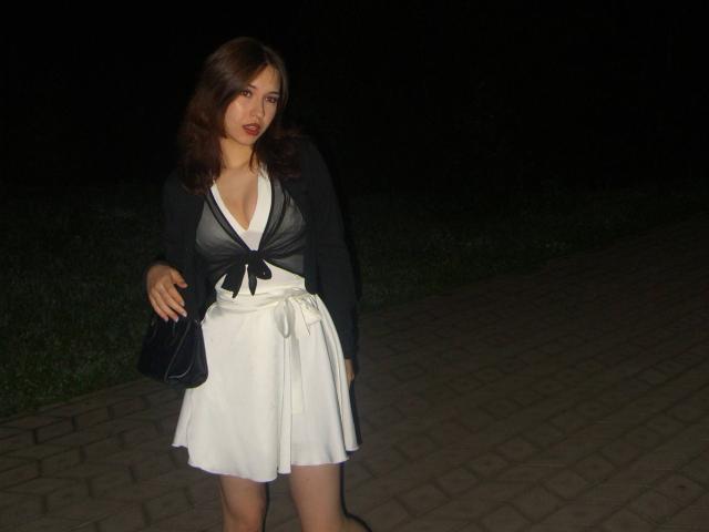 AletaBreslauer has brown wavy hair, red lipstick, black cardigan, white dress with black top, and black purse, standing on brick pavement at night, showing her tits slightly.