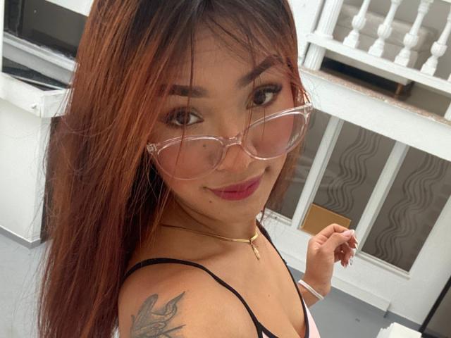 SashaHans with long brown hair, pink glasses, and pink lipstick, showing a tattoo on her shoulder, wearing a black top, smiling slightly, holding her hair.