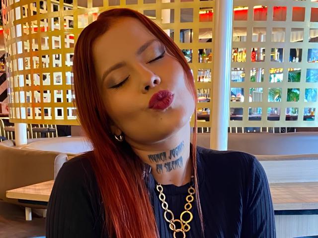 ElectraRoss with red hair, eyes closed, pouting lips, black top, gold necklace, tattoo under chin, and small hoop earrings, in a brightly lit room.
